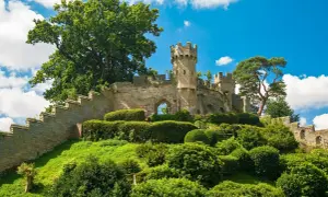Warwick Castle reser sig stolt på en grönskande kulle, omgiven av frodig grönska och en klarblå himmel med vita moln.