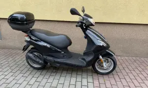 Czarny skuter Piaggio Fly 50 z kuferkiem na bagaż, zaparkowany na chodniku z kostki brukowej.