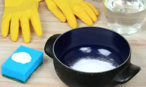 Gants jaunes, éponge bleue avec bicarbonate, casserole noire et un bocal de liquide. Astuces pour savoir comment nettoyer une casserole qui a brûlé.