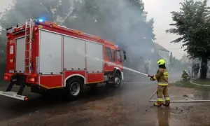 Strażak OSP w akcji, z węża wodnego leje wodę na tle remizy i drzew.