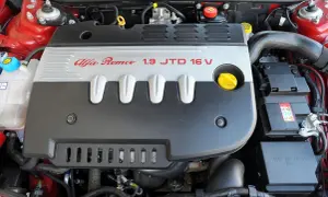 Silnik Alfa Romeo 147 1.9 JTD 16V, widok z góry. Czerwona karoseria, czarna osłona silnika z napisem.