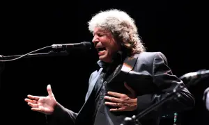 Un cantante de grupos flamencos de los 80, con melena rubia, canta apasionadamente frente a un micrófono, con la mano en el pecho.