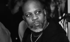 DMX, z siwą brodą i łańcuchem na szyi, spogląda w bok. W tle widać niewyraźne sylwetki, być może jego dzieci.