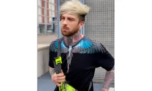 Hombre con tatuajes visibles en cuello y brazos, viste camiseta negra con alas azules y bolso neón.