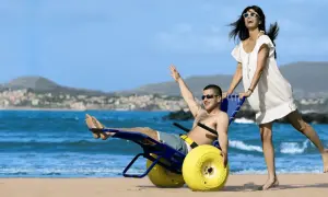 Coppia felice sulla spiaggia, lui su una sedia job con ruote gialle, lei lo spinge. Mare e collina sullo sfondo.