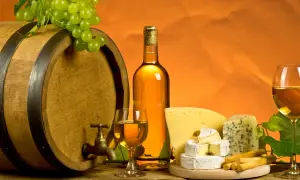 Drewniana beczka, butelka i kieliszki białego wina, winogrona, różne sery i paluszki na drewnianym stole.