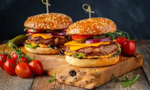 Dwa soczyste burgery z idealnie wysmażonym mięsem na burgery, serem, warzywami i sezamową bułką, na drewnianej desce.