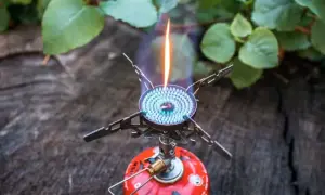 Soto Windmaster Kocher mit blauer Flamme auf einem Baumstamm, umgeben von grünen Blättern.