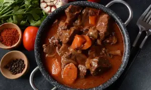 Gulasz z sarny po myśliwsku, aromatyczny, z kawałkami mięsa, marchewką i cebulą w gęstym sosie. Obok przyprawy i warzywa.