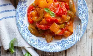 Piatto di peperonata con patate, colorata e saporita, guarnita con basilico fresco su un piatto decorato.