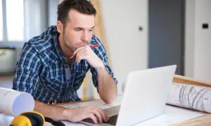 Mężczyzna w kratę na laptopie, z ołówkiem w dłoni, szuka informacji, jak sprawdzić wiarygodność firmy. Obok leżą plany i słuchawki.