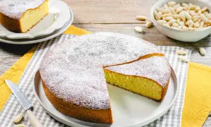 Torta soffice con farina di mandorle, spolverata di zucchero a velo. Accanto, una ciotola di mandorle intere e una fetta già servita.
