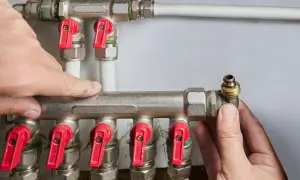 Instalator montuje podłączenie dolne grzejnika PEX. Widać rozdzielacz z czerwonymi zaworami i białe rury.