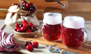Due boccali di birra kriek rossastra con schiuma bianca, circondati da ciliegie fresche su un tagliere di legno.