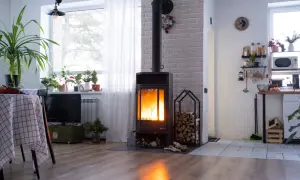 Płonący kominek z podłączeniem do komina systemowego, obok stos drewna. W tle kuchnia i okno z roślinami.