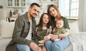 Szczęśliwa rodzina z dwójką dzieci siedzi na kanapie. Rodzice i dzieci uśmiechają się. Program 800 plus obejmuje dzieci do 18 roku życia.