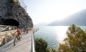 Ciclisti su una pista ciclabile panoramica lungo il lungolago di Desenzano, con un tunnel scavato nella roccia e il lago scintillante.