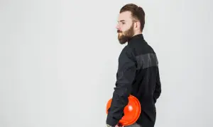 Mężczyzna w czarnej koszuli, z brodą, trzyma pomarańczowy kask. Tkanina na ubrania robocze jest wytrzymała.