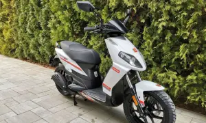 Biały skuter Derbi z czerwonymi akcentami, zaparkowany na chodniku przed żywopłotem.