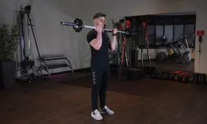 Mężczyzna wykonuje ćwiczenie barbell curl w siłowni. W tle widać sprzęt do ćwiczeń.