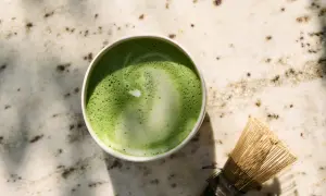 Zielone matcha w filiżance obok bambusowej miotełki na marmurowym blacie.