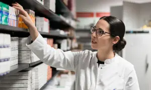 Farmaceutka w białym kitlu sięga po lek, być może pentoksyfilinę, z aptecznej półki pełnej opakowań.