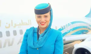 Uśmiechnięta stewardessa w niebieskim mundurku i charakterystycznym nakryciu głowy pozuje przed samolotem.