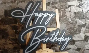 Czarny napis "Happy Birthday" z białymi obrysami, stojący na sztaludze. Idealny na życzenia "sto lat"!