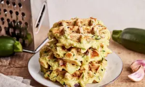 Stapel goldbrauner Zucchini-Waffeln auf einem Teller, daneben Zucchini, Knoblauch und eine Reibe.