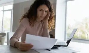 Zmartwiona kobieta analizuje dokumenty, rozważając odrzucenie spadku. Na stole leży laptop i kalkulator.