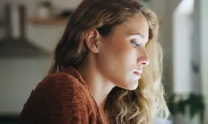 Młoda kobieta z blond włosami, w brązowym swetrze, patrzy w dal. Jej wyraz twarzy sugeruje dysocjację, jakby była nieobecna myślami.