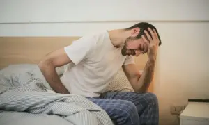 Mann sitzt im Bett, hält sich den Kopf. Mögliche Bewegungsmangel Symptome wie Kopfschmerzen und Müdigkeit sind zu erkennen.
