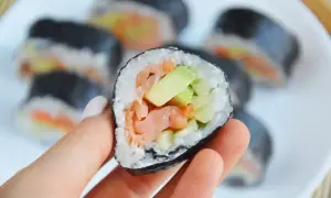 Smaczny futomak z łososiem i awokado, trzymany w dłoni na talerzu pełnym sushi.