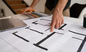 Dłoń wskazująca na plan mieszkania, obok laptop i próbki materiałów.