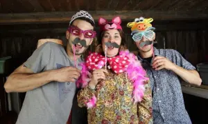 Tres amigos se divierten con disfraces hechos con cosas de casa: un hombre con tiara y bigote, una mujer con orejas de gato y un hombre con máscara de vaca.