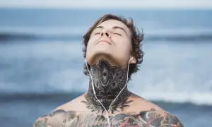 Mężczyzna z tatuażem na szyi i klatce piersiowej, słuchający muzyki na plaży. Jego oczy są zamknięte, a głowa lekko uniesiona.