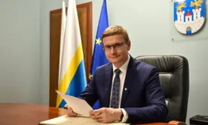 Mężczyzna w garniturze siedzi przy biurku, trzymając dokumenty. Za nim flagi i herb miasta, symbolizujące jego rolę w obszarze prawo administracyjne.