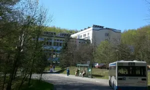 Sanatorium uzdrowiskowe „Leśnik” otoczone zielenią, z autobusem na pierwszym planie i ludźmi czekającymi na przystanku.