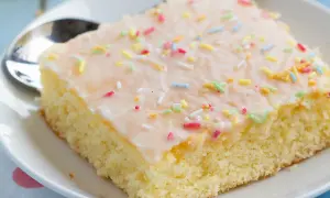Ein Stück saftiger Fantakuchen vom Blech, mit Zuckerguss und bunten Streuseln bestreut, auf einem Teller mit Löffel.