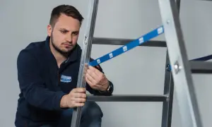 Mężczyzna w granatowej bluzie z logo KRAUSE przeprowadza badanie drabin, sprawdzając mocowanie niebieskiej taśmy zabezpieczającej.