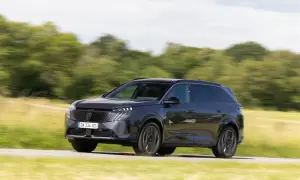 Ciemnoszary Peugeot 5008 pędzi po drodze, otoczony zielenią pól i drzew.