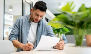Giovane uomo concentrato su documenti, con una pianta rigogliosa che suggerisce un ambiente di lavoro moderno e un uso foresteria.