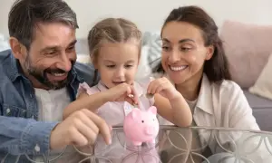 Rodzina z uśmiechem wrzuca monety do świnki-skarbonki. Dziecko uczy się oszczędzać, by zebrać 12 tys na dziecko.