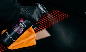 Ręka w czarnej rękawiczce rozpyla quick detailer K2 Spectrum Pro na czarną powierzchnię z czerwonym wzorem. Pomarańczowa mikrofibra obok.