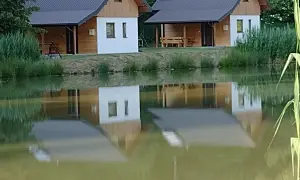 Urokliwe łowisko z domkami w Podkarpackiem. Dwa drewniano-białe domki z dachami odbijają się w spokojnej tafli wody.