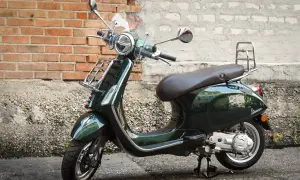 Elegancki, ciemnozielony skuter Piaggio z brązowym siedzeniem i chromowanymi dodatkami stoi na tle ceglanej ściany.