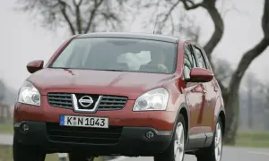 Czerwony Nissan Qashqai 1.6 benzyna, często chwalony w opiniach, jedzie po drodze wśród drzew.