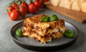 En bit italiensk lasagne med köttfärssås och ost, garnerad med basilika. Tomater och parmesanost i bakgrunden.