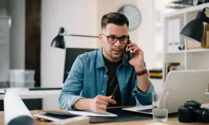 Mężczyzna w okularach rozmawia przez telefon przy biurku z laptopem i aparatem.