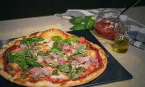 En läcker spenatpizza med prosciutto, basilika och pesto. Bredvid står en burk tomatsås och en flaska olivolja.